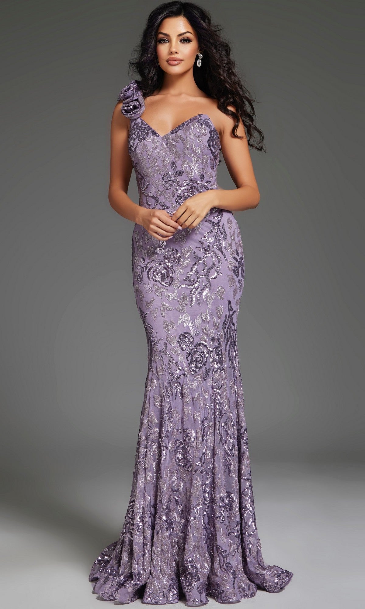 A woman with wavy dark hair wears the Jovani 37634 long lavender prom dress, featuring sequins, floral patterns, and a single shoulder strap adorned with a fabric flower.